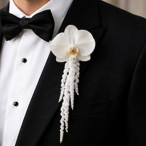 May include: A white orchid boutonniere featuring a single large bloom with a yellow center and cascading white astilbe flowers, pinned to the lapel of a black suit jacket worn with a white dress shirt and a black bow tie.