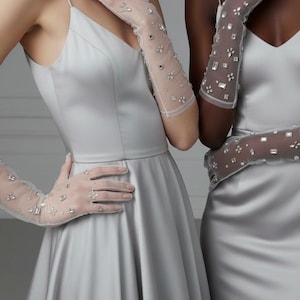 Può includere: Due donne indossano abiti di raso argentato e lunghi guanti trasparenti ornati di cristalli scintillanti. I guanti arrivano fino alla parte superiore del braccio, creando un look elegante e glamour. Gli abiti hanno un design semplice e classico.