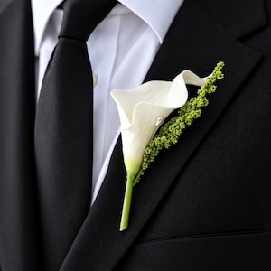 Op de afbeelding: Een witte calla lelie corsage met een groene steel en een textuurrijke groene vulbloem, vastgezet met een kleine speld met parelkop, opgespeld op de revers van een zwart colbert gedragen over een wit overhemd en een zwarte stropdas.
