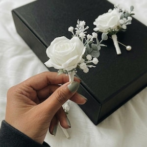 May include: Two white rose boutonnieres with greenery and pearl accents. The boutonnieres are on a black box, ideal for a wedding. The roses are the focal point, with delicate leaves and a pearl pin.