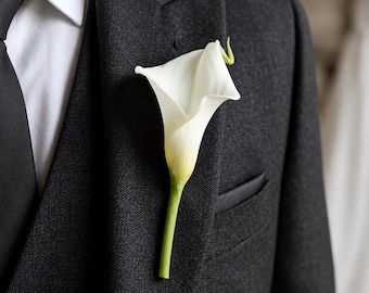 Può includere: Un singolo fiore all'occhiello a forma di calla bianca appuntato sul bavero di una giacca da abito in lana grigio antracite scuro. L'abito è indossato con una camicia bianca e una cravatta nera tinta unita, per un aspetto formale e curato.