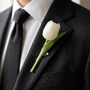 May include: A single white tulip boutonniere with a green stem and leaf, secured to the lapel of a dark charcoal gray wool blazer with a small silver pin featuring a white pearl accent, worn over a crisp white collared shirt and black tie.