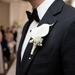 Op de afbeelding: Een witte calla lelie corsage met kleine witte vulbloemen en een wit lint, opgespeld op de revers van een zwart colbert gedragen over een wit overhemd met een zwarte vlinderdas, voorzien van een klein parelaccent aan de onderzijde.