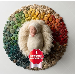 May include: A newborn baby swaddled in cream-coloured fabric and a matching hat, resting in a fluffy, cream-coloured nest. The nest is encircled by a ring of colourful buttons, with a red banner reading "1 Newborn Backdrop."