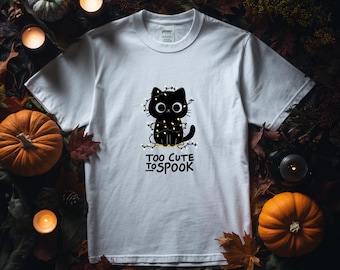 Op de afbeelding: Wit T-shirt met een zwarte kattenillustratie omwikkeld met lichtsnoeren. De kat heeft grote, ronde ogen en zit. De tekst "TOO CUTE TO SPOOK" staat eronder. Het shirt is omgeven door pompoenen, bladeren en kaarsen.