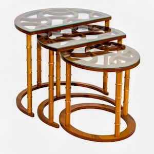 May include: Set of three nesting tables with a unique design. Each table has a clear glass top with a decorative wooden base. The legs are bamboo-style, and the tables are arranged in a descending size order.