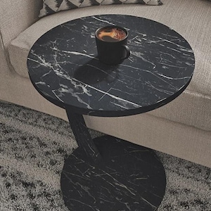 May include: A black marble-effect side table with a single curved leg and a round base. A black coffee mug with coffee sits on the table. The table is next to a beige sofa with a patterned cushion.