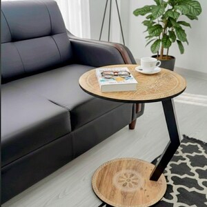 May include: A wooden side table with a carved design on the top and base. The table has a black metal support. A book, glasses, and a teacup sit on the table. The table is next to a black leather sofa.