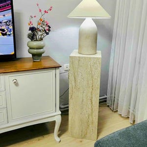 May include: A beige table lamp with a cream-colored lampshade sits atop a rectangular stone pedestal. The lamp has a rounded base and a cylindrical body. A vase with colorful flowers is on a wooden cabinet.