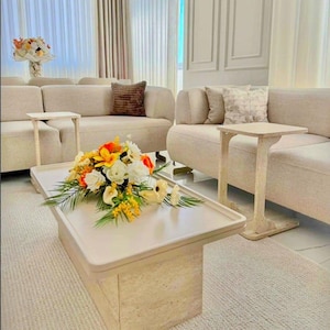 May include: A contemporary living room setting with beige sofas, a rectangular coffee table, and side tables. The coffee table displays a floral arrangement with white, yellow, and orange flowers. The room has neutral walls and a light rug.