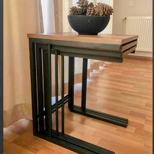 Puede incluir: Un juego de dos mesas nido con un diseño industrial moderno. Las mesas tienen una estructura de metal oscuro y una tapa de madera. La mesa superior mide 61 cm de largo, 41 cm de ancho y 51 cm de alto. Las mesas están rematadas con un cuenco negro lleno de piñas.