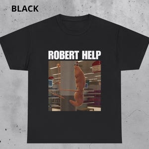 Könnte beinhalten: Schwarzes T-Shirt mit dem Text "ROBERT HELP" in weißen Blockbuchstaben. Das Shirt zeigt eine humorvolle Grafik einer garnelenartigen Kreatur, die in einem Lebensmittelgeschäft hängt. Das Wort "BLACK" ist oben aufgedruckt.