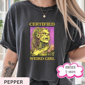 May include: Dark gray unisex t-shirt with the text "CERTIFIED WEIRD GIRL" above and below a graphic of a green-skinned character with yellow hair and purple background. The shirt also has the word "PEPPER" and "UNISEX T-shirt" on it.