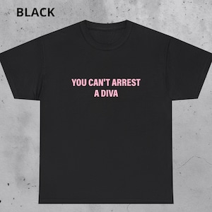 Puede incluir: Camiseta negra con el texto rosa "YOU CAN'T ARREST A DIVA". La palabra "BLACK" está impresa en la esquina superior izquierda. Una prenda de vestir informal.