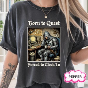 May include: Dark grey t-shirt with a distressed finish. The graphic shows a knight in armour at a desk with a computer, a sword, and the text "Born to Quest, Forced to Clock In". A pink cloud-shaped logo with the word "PEPPER" is in the bottom right corner.