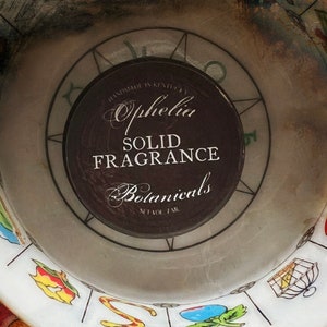 Op de afbeelding: Een witte keramische schaal met een kleurrijk ontwerp rond de rand. In het midden van de schaal bevindt zich een zwart label met de tekst "Ophelia Solid Fragrance Botanicals".