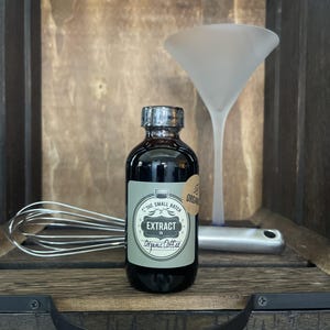May include: A dark glass bottle of "Extract Organic Coffee" with a silver cap, a label, and a small round sticker. A frosted martini glass and a whisk are in the background, all set in a wooden crate.