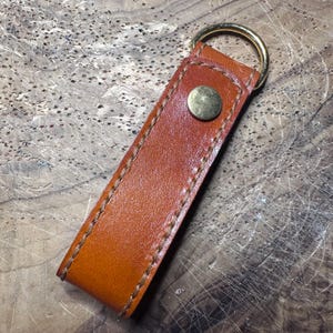 May include: A rectangular tan leather keyring featuring a smooth texture and visible brown stitching along the edges. The leather is folded over a circular gold-toned metal ring and secured with a single round gold-toned metal snap button fastener.