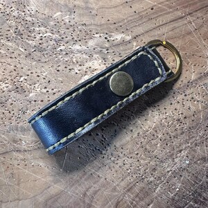 May include: A black leather keychain strap featuring a brass-toned metal snap button closure and a circular brass key ring. The strap is constructed with visible light-coloured stitching along the edges, securing the folded leather loop together.