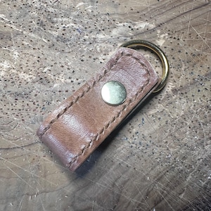 May include: A brown leather keyring featuring a rectangular folded strap with visible stitching along the edges. A smooth, circular brass-toned metal snap button is centred on the leather, and a gold-toned metal split ring is attached at the top.