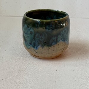 Puede incluir: Una taza de cerámica hecha a mano con forma redondeada. La taza presenta un esmalte verde oscuro y azul que gotea sobre una base beige. El interior de la taza es oscuro y el borde es liso.