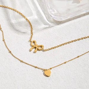 Könnte beinhalten: Eine mehrreihige, goldfarbene Halskette mit zwei Ketten. Die obere Kette hat einen Anhänger in Schleifenform, während die untere Kette einen herzförmigen Anhänger hat. Die Halskette wird auf einer hellen Oberfläche gezeigt.
