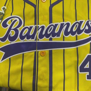May include: A yellow baseball jersey with vertical purple pinstripes, featuring the word Bananas in large purple script across the chest, a purple number 4 on the lower right, and a circular team logo patch on the upper left shoulder area.