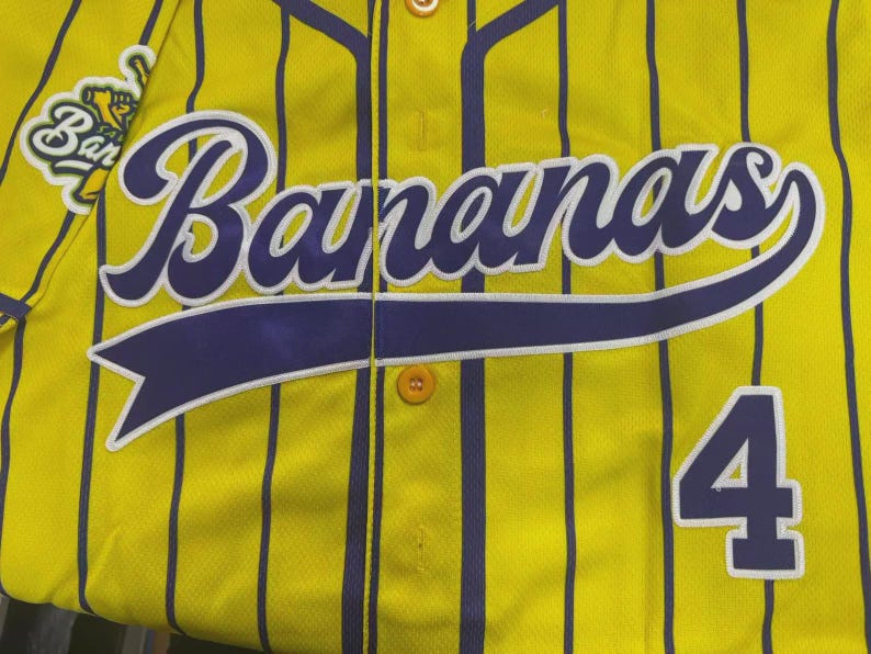 May include: A yellow baseball jersey with vertical purple pinstripes, featuring the word Bananas in large purple script across the chest, a purple number 4 on the lower right, and a circular team logo patch on the upper left shoulder area.