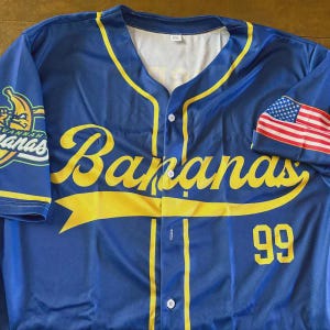 May include: A blue button-down baseball jersey featuring the yellow script text Bananas across the chest, a yellow banana logo on the left sleeve, the number 99 in yellow on the front, and an American flag graphic on the right sleeve.
