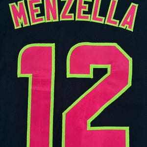 May include: A black jersey featuring the name MENZELLA in hot pink letters with a lime green outline, positioned above the number 12, which is also rendered in hot pink with a lime green outline. The fabric has a visible mesh texture.