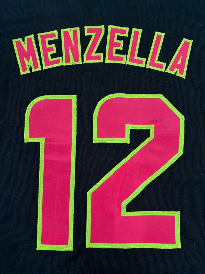 May include: A black jersey featuring the name MENZELLA in hot pink letters with a lime green outline, positioned above the number 12, which is also rendered in hot pink with a lime green outline. The fabric has a visible mesh texture.