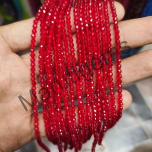 Puede incluir: Hebras de pequeñas cuentas rojas facetadas. Las cuentas están ensartadas, creando múltiples líneas verticales. Las cuentas tienen una superficie brillante y reflectante. La imagen es un primer plano, que muestra la textura y el color de las cuentas. Las cuentas son probablemente para hacer joyas.