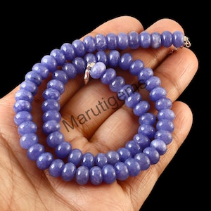 Puede incluir: Un collar de cuentas redondas y pulidas, de color azul lavanda. Las cuentas están ensartadas con un cierre plateado. El collar se muestra sobre un fondo oscuro.