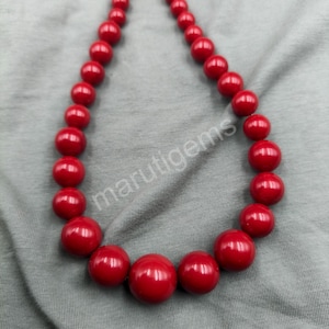 Puede incluir: Un collar con grandes cuentas rojas brillantes. Las cuentas están ensartadas, formando una forma circular. El collar se muestra sobre una superficie gris. El texto "marutigems" es visible.