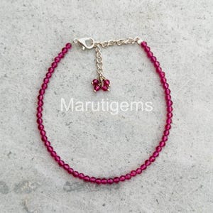 Peut inclure: Un bracelet délicat, composé de petites pierres précieuses rondes et rouge foncé. Le bracelet est doté d'une chaîne et d'un fermoir dorés, avec un petit groupe de pierres précieuses assorties suspendues à la chaîne. Le texte "Marutigems" est visible.