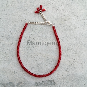 May include: A delicate red beaded bracelet with a silver chain and clasp. The bracelet features small, round beads and a cluster of beads at the end of the extender chain. The brand name "Marutigem" is visible on the background.