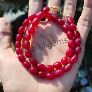 Puede incluir: Un collar de dos hilos con cuentas ovaladas de color rojo intenso. El collar se sostiene en una mano, con un cierre dorado visible. El texto "arutigems" es parcialmente visible en la mano.