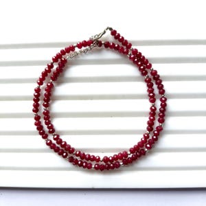 May include: A double-strand necklace featuring small, faceted, deep red beads. The necklace has a silver-colored clasp and is displayed on a white surface. The beads are evenly spaced, creating a delicate and elegant jewelry piece.