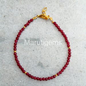 Puede incluir: Una pulsera delicada con pequeñas cuentas facetadas de color rojo intenso dispuestas en forma circular, acentuada con diminutos espaciadores dorados y un cierre de mosquetón dorado. El texto Marutigems está centrado dentro de la pulsera.