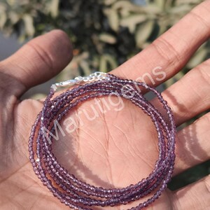 Könnte beinhalten: Eine mehrsträngige Halskette mit kleinen, facettierten, tiefvioletten Edelsteinperlen. Die Kette verfügt über einen silberfarbenen Metallverschluss und eine Verlängerungskette. Der Text Marutigems ist über die violetten Perlenstränge gelegt.