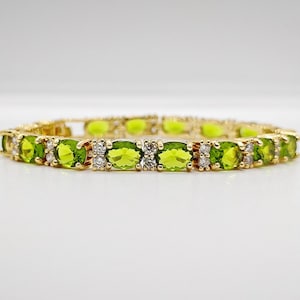 May include: A gold-toned tennis bracelet featuring a repeating pattern of oval-cut lime green peridot gemstones alternating with pairs of small, clear, round-cut white stones set in a metallic frame that encircles the wrist in a continuous line.