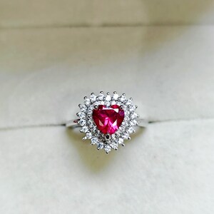 Puede incluir: Un anillo de plata con una gema roja en forma de corazón, rodeada por un halo de piedras claras más pequeñas. El diseño del anillo es elegante y llamativo, ideal para ocasiones especiales o uso diario. El anillo se encuentra sobre un fondo suave y neutro.