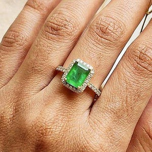 Könnte beinhalten: Ein goldener Ring mit einem rechteckigen, smaragdgeschliffenen grünen Edelstein, der von einem Kranz aus kleinen weißen Steinen umgeben ist. Die Ringschiene ist schmal und ebenfalls in der oberen Hälfte mit kleinen weißen Steinen besetzt.