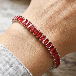 Può includere: Un bracciale tennis caratterizzato da una fila continua di pietre preziose rosse rettangolari a taglio baguette, incastonate in una montatura in metallo color argento. Il bracciale avvolge il polso, mostrando una vivace tonalità cremisi.
