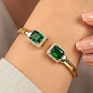 May include: A gold-colored cuff bracelet with two emerald-cut green gemstones. Each gemstone is surrounded by a halo of small, clear stones. The bracelet is open and worn on a wrist.