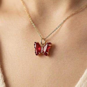 Peut inclure: Un collier doré avec un pendentif papillon. Le pendentif papillon est rouge vif, encadré d'or et orné de petits détails blancs. Le collier est porté autour du cou.