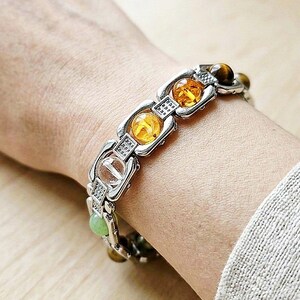 Puede incluir: Una pulsera de plata con una serie de gemas redondas y coloridas. La pulsera presenta piedras ámbar, claras y verdes, conectadas por eslabones plateados ornamentados. La pulsera se lleva en la muñeca, mostrando el diseño y los colores de la joya.