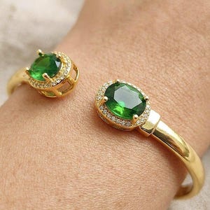 Peut inclure: Un bracelet manchette ouvert doré orné de deux pierres ovales vert émeraude, chacune entourée d'un halo de petits cristaux transparents pavés. Le bracelet présente une bande métallique lisse et polie, ouverte au centre pour un ajustement flexible.