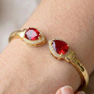 Può includere: Un bracciale aperto color oro con due pietre sfaccettate rosso intenso a forma di pera su ciascuna estremità. Ogni pietra rossa è circondata da un alone di piccoli cristalli trasparenti incastonati a pavé su una fascia in metallo dorato lucido.