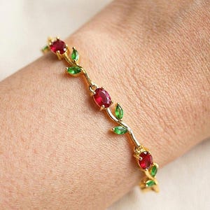 Könnte beinhalten: Ein goldfarbenes Armband mit einem rankenartigen Design, das abwechselnd mit oval geschliffenen roten Edelsteinen und marquiseförmig geschliffenen grünen Edelsteinen besetzt ist. Die Metallglieder verbinden die Steine zu einer flexiblen Kette.